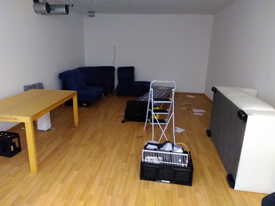 Des Chaostreff Berns Hackcenter. Ein leerer Holztisch zur linken, ein teilweise abgebautes Sofa in der Ecke, ein weiteres Sofa zur Rechten liegt auf dem Rücken. Mitten im Raum steht eine Leiter.