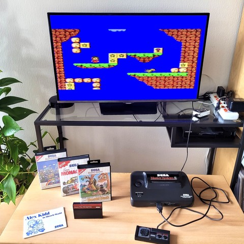 A SEGA Master System II, with the games 'The Lucky Dime Caper,' 'Rainbow Islands,' 'Super Monaco GP II,' and 'Sonic the Hedgehog.' On the TV screen: 'Alex Kidd in Miracle World.'