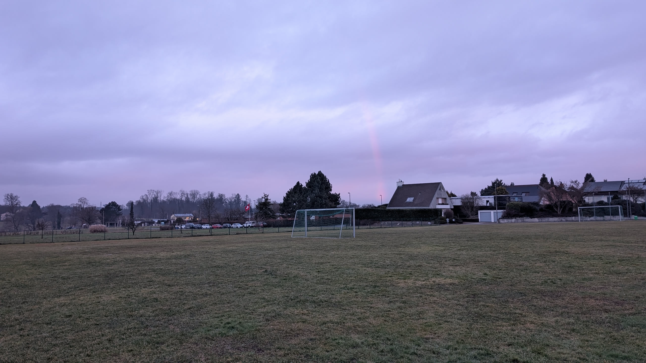 Photo of the view across a grass pitch and some houses under a grey sky. Right in the centre, there is a piece of a red line that looks very much like a rainbow, just red.